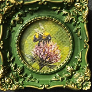 May include: A small square painting in a decorative green and gold frame featuring a bumblebee perched on a pink clover flower. The artwork uses textured brushstrokes against a light green background, capturing the insect's yellow and black body.