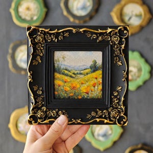 K&ouml;nnte beinhalten: Ein quadratisches Gem&auml;lde eines gelben Mohnfeldes unter blauem Himmel, eingefasst in einen schwarzen Rahmen mit kunstvollen, goldfarbenen Rosenreliefs. Das Kunstwerk zeigt eine h&uuml;gelige Landschaft mit fernen Bergen und gr&uuml;nem Blattwerk.