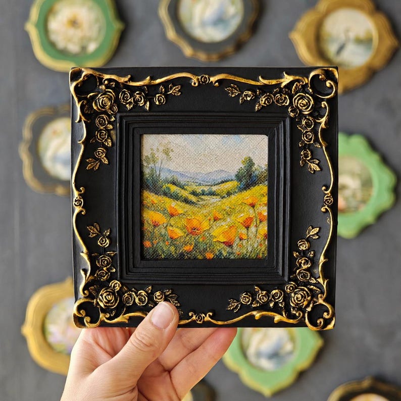 K&ouml;nnte beinhalten: Ein quadratisches Gem&auml;lde eines gelben Mohnfeldes unter blauem Himmel, eingefasst in einen schwarzen Rahmen mit kunstvollen, goldfarbenen Rosenreliefs. Das Kunstwerk zeigt eine h&uuml;gelige Landschaft mit fernen Bergen und gr&uuml;nem Blattwerk.