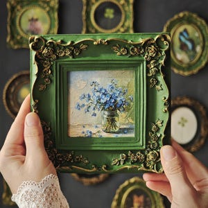 May include: A square painting of blue flowers in a glass jar, encased in a dark green frame with ornate gold-painted rose details. The frame features a textured, raised floral relief pattern along its edges, surrounding the central canvas.