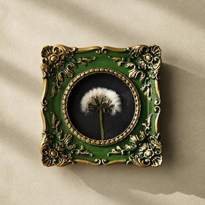 May include: A square wall frame featuring an ornate green and gold border with intricate scrollwork. The frame contains a circular black insert displaying a single white dandelion seed head with a thin green stem centered against the dark background.