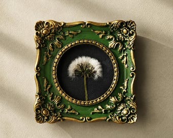 May include: A square wall frame featuring an ornate green and gold border with intricate scrollwork. The frame contains a circular black insert displaying a single white dandelion seed head with a thin green stem centred against the dark background.