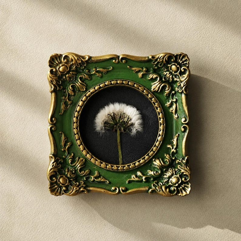 May include: A square wall frame featuring an ornate green and gold border with intricate scrollwork. The frame contains a circular black insert displaying a single white dandelion seed head with a thin green stem centred against the dark background.