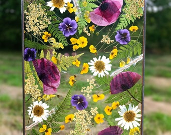 May include: A framed pressed flower art piece with a variety of flowers, including purple pansies, white daisies, yellow wildflowers, and pink petals. The flowers are pressed between two pieces of glass and framed in a black metal frame.