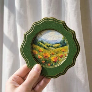 May include: A small, round, green frame with a gold trim, holding a miniature painting of a field of orange flowers and rolling hills. The frame has a scalloped edge and is held up by a hand.