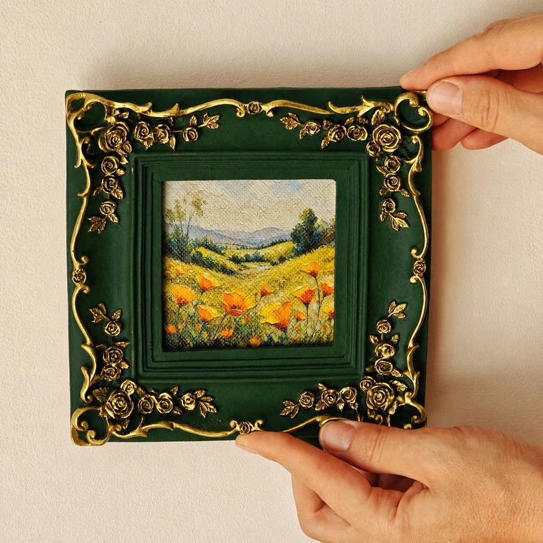 K&ouml;nnte beinhalten: Ein quadratisches Gem&auml;lde in einem dunkelgr&uuml;nen Rahmen mit verzierten goldfarbenen Rosenapplikationen. Das Kunstwerk zeigt eine Landschaft mit sanften H&uuml;geln, einer Wiese und leuchtend orangefarbenen Blumen unter einem weichen Himmel.