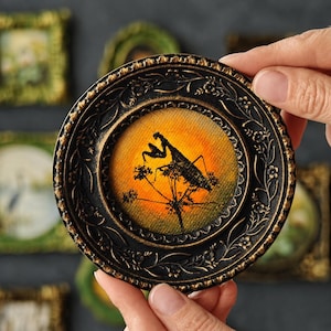May include: A small circular wall art piece featuring a black silhouette of a praying mantis perched on a plant against a vibrant orange and yellow sunset background, encased in an ornate black frame with embossed floral and vine details.