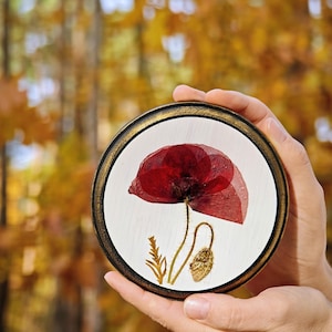 May include: A circular artwork with a pressed red poppy flower, set against a white backdrop and framed in gold. The artwork is held in hands, with an out-of-focus autumnal background of yellow and brown leaves.