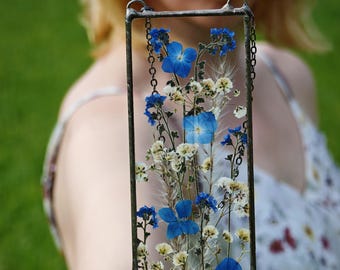 May include: A rectangular glass and metal frame filled with dried blue and white flowers. The frame has a chain for hanging. The flowers are a mix of blue blooms and delicate white blossoms, creating a natural, artistic display.