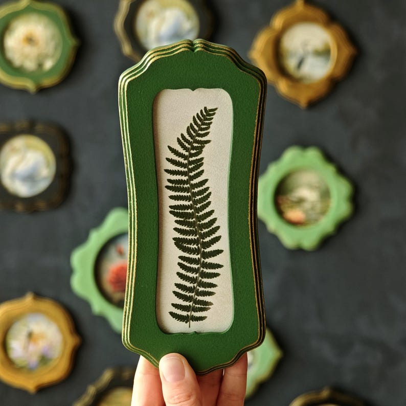 Puede incluir: Un marco de pared alto y rectangular con un acabado verde oscuro y bordes dorados. El marco contiene un fondo de color claro con una ilustraci&oacute;n de una hoja de helecho verde oscuro. El marco tiene un dise&ntilde;o decorativo de bordes festoneados.