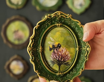 Può includere: Un dipinto a olio ovale raffigurante un bombo su un fiore di trifoglio rosa, racchiuso in una cornice verde scuro ornata con riflessi dorati. Il dipinto presenta pennellate strutturate e uno sfondo verde vivace con l'insetto dettagliato.