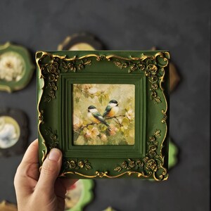 May include: A square wall art piece featuring a painting of two small birds perched on a flowering branch. The artwork is encased in a dark green frame with ornate, raised gold-coloured rose and leaf detailing along the edges and corners.