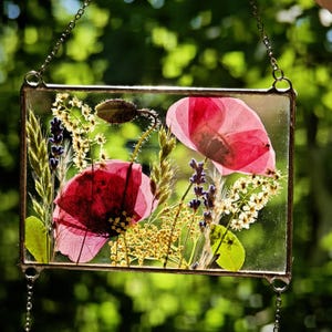 May include: A rectangular glass art piece featuring pressed red poppies and other dried flowers. The artwork is encased in a metal frame and suspended by a chain, showcasing a natural, botanical theme.