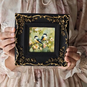 May include: A square painting of two small birds perched on a flowering branch, set within a black frame featuring ornate gold-coloured rose and vine relief detailing along the edges. The artwork depicts a soft, green, and floral background.