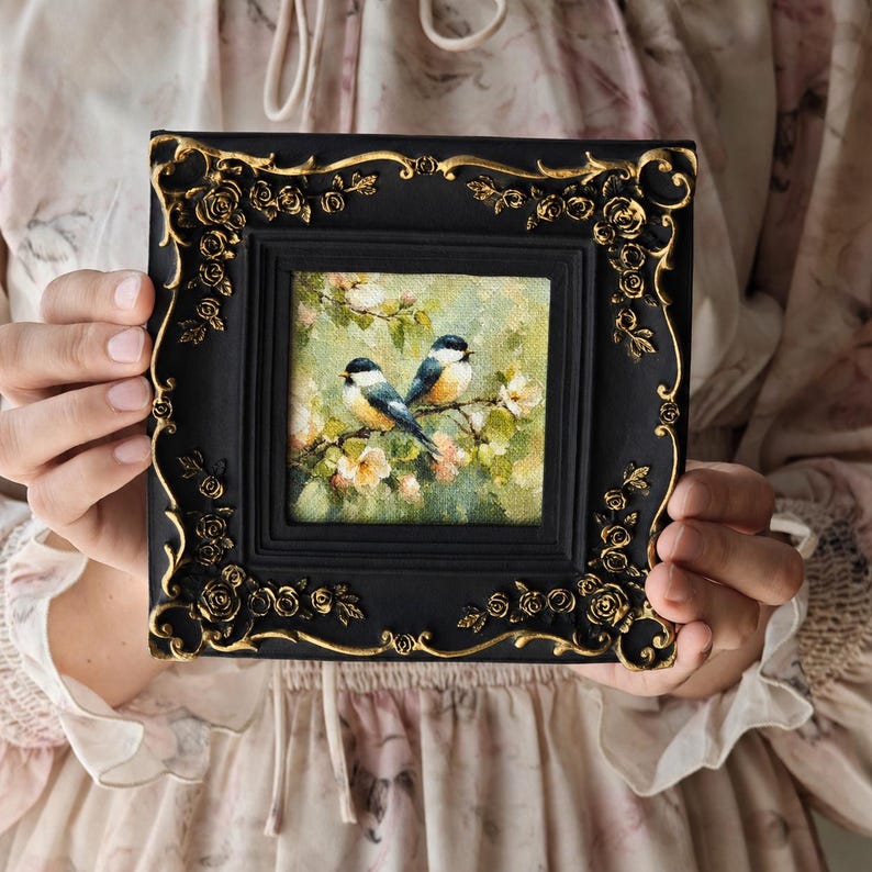 May include: A square painting of two small birds perched on a flowering branch, set within a black frame featuring ornate gold-coloured rose and vine relief detailing along the edges. The artwork depicts a soft, green, and floral background.