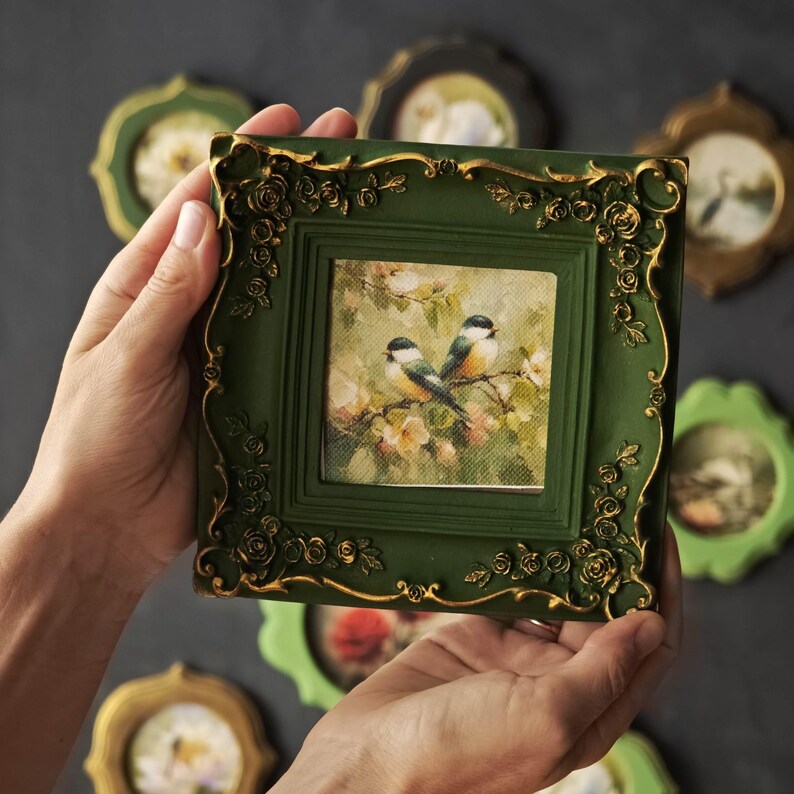 May include: A square picture frame in dark green with gold-coloured floral relief detailing. The frame holds a print of two small birds perched on flowering branches. The frame features ornate, raised rose motifs at the corners and edges.