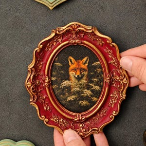 May include: An oval-shaped wall art piece featuring a portrait of a fox surrounded by foliage. The frame is deep red with ornate gold-painted floral and scrollwork detailing along the edges. The central image is set against a dark background.