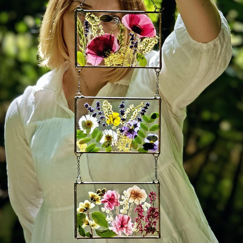 May include: A three-panel hanging art piece featuring pressed flowers and foliage. Each rectangular glass panel is framed in metal and connected by chains. The top panel displays red poppies, the middle panel has yellow and purple flowers, and the bottom panel showcases pink and white blooms.