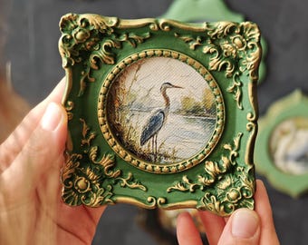 May include: A small square frame with ornate gold and green detailing surrounding a circular painting of a heron standing by a body of water. The frame features intricate scrollwork and relief patterns at the corners and edges.