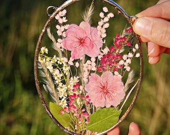 May include: An oval-shaped glass and metal frame with pressed flowers. The arrangement includes pink flowers, white and red accents, and green leaves. The frame is held up by a chain and is surrounded by a silver metal border.