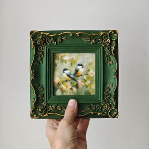 May include: A square painting of two small birds perched on a flowering branch, encased in a dark green frame with ornate gold-coloured floral relief detailing along the edges. The artwork features soft brushstrokes and a light, natural colour palette.