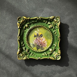May include: A small square picture frame in dark green with gold-toned ornate detailing, enclosing a circular oil painting of a bumblebee perched on a pink clover flower against a soft yellow and green background.