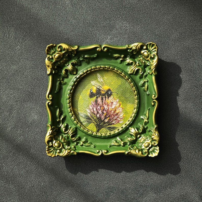 May include: A small square picture frame in dark green with gold-toned ornate detailing, enclosing a circular oil painting of a bumblebee perched on a pink clover flower against a soft yellow and green background.