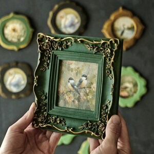 May include: A square picture frame in dark green with gold-painted floral and rose relief detailing at the corners. The frame holds a print of two small birds perched on branches amidst soft-toned flowers, set against a dark background.