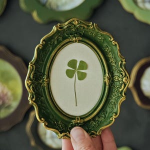 May include: An oval-shaped wall frame with a dark green finish and gold-toned highlights along the ornate, scalloped edges. The frame contains a light-colored backing featuring a single, pressed four-leaf clover centered within the display area.