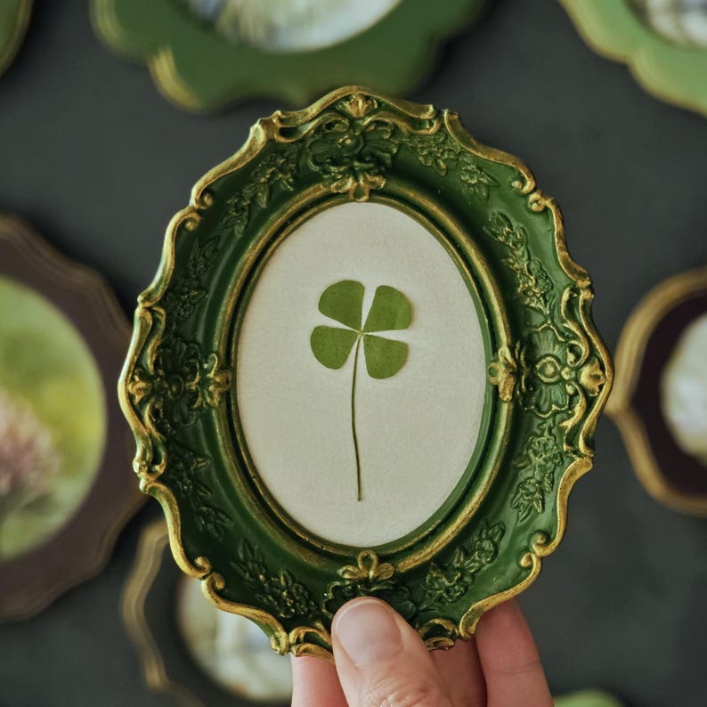 May include: An oval-shaped wall frame with a dark green finish and gold-toned highlights along the ornate, scalloped edges. The frame contains a light-colored backing featuring a single, pressed four-leaf clover centered within the display area.