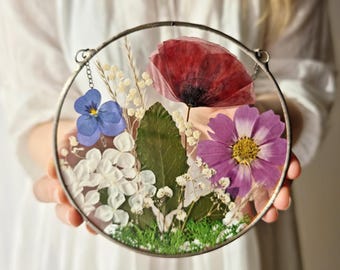 May include: A round glass frame with a silver metal border, containing pressed flowers. The flowers include a red poppy, a purple cosmos, a blue pansy, and white flowers. The frame is hanging from a silver chain.
