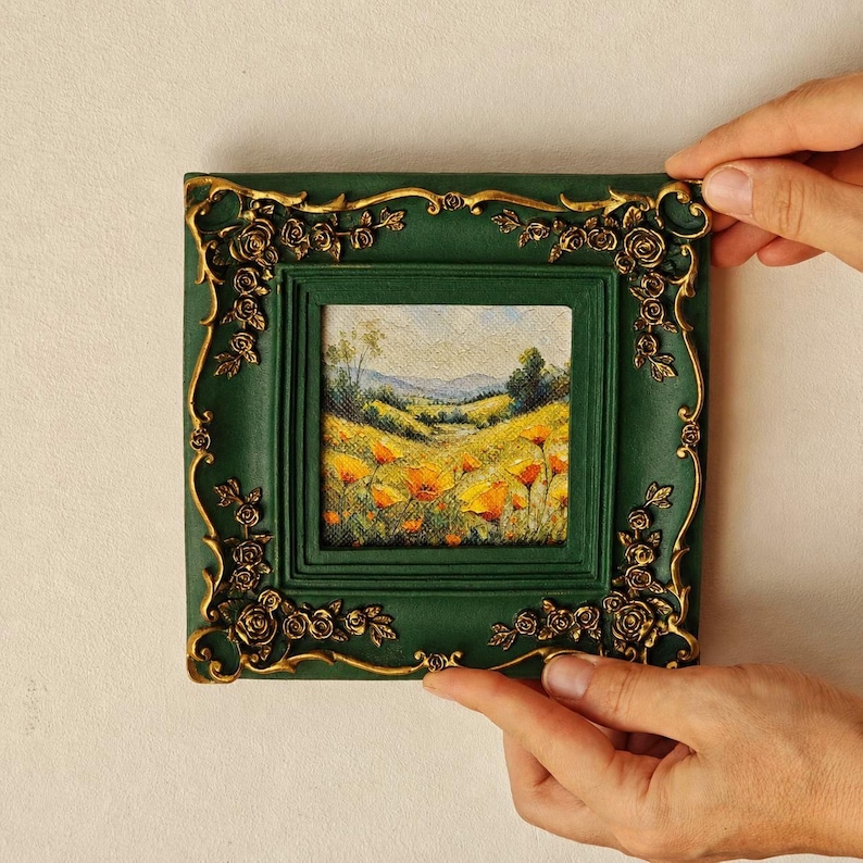 K&ouml;nnte beinhalten: Ein quadratischer, dunkelgr&uuml;ner Bilderrahmen mit verzierten, goldfarbenen floralen Reliefdetails an den Ecken. Der Rahmen umschlie&szlig;t ein kleines Landschaftsgem&auml;lde, das ein leuchtendes Feld mit gelben und orangen Blumen zeigt.