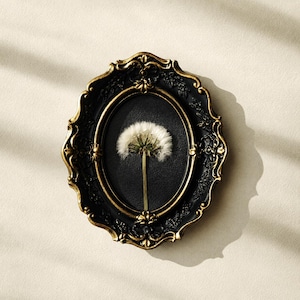 May include: An oval wall frame with a black and gold finish featuring ornate floral carvings. The frame contains a black background with a single white dandelion seed head centered inside, displaying a delicate stem and fluffy white seeds.