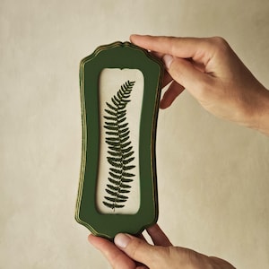 Puede incluir: Un marco de pared rectangular vertical con acabado verde oscuro y bordes dorados. El marco presenta un borde festoneado y contiene una inserci&oacute;n de color blanco roto que muestra una ilustraci&oacute;n detallada de una hoja de helecho verde oscuro.