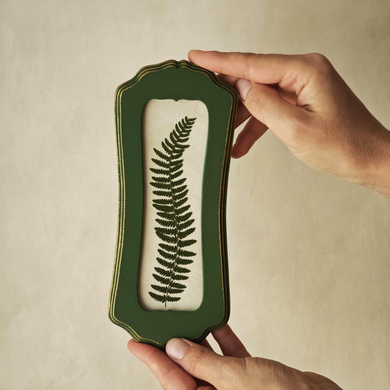 Puede incluir: Un marco de pared rectangular vertical con acabado verde oscuro y bordes dorados. El marco presenta un borde festoneado y contiene una inserci&oacute;n de color blanco roto que muestra una ilustraci&oacute;n detallada de una hoja de helecho verde oscuro.