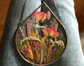 May include: A teardrop-shaped glass ornament with dried flowers and foliage pressed inside. The ornament is hung from a silver chain and features pink, red, and yellow flowers.