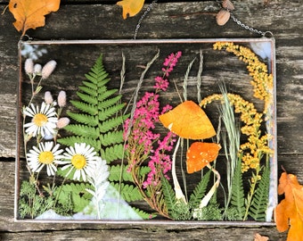 Könnte beinhalten: Ein rechteckiges Glaskunstwerk mit gepressten Blumen und Pflanzen. Die Anordnung umfasst weiße Gänseblümchen, grüne Farne, rosa Blüten, orangefarbene Pilze und gelbe Wildblumen. Es ist mit einem silbernen Metallrahmen eingefasst und an einer Kette aufgehängt.