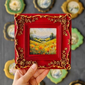 K&ouml;nnte beinhalten: Ein quadratisches Gem&auml;lde in einem tiefroten Rahmen mit verzierten goldfarbenen Rosenverzierungen an den Ecken. Das Kunstwerk zeigt eine lebendige Landschaft mit einem Feld aus gelben und orangefarbenen Wildblumen unter einem sanften Himmel.