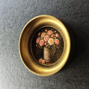 May include: A miniature painting of a floral arrangement, featuring pink and cream roses in a metallic jug, presented in a gold-coloured circular frame. The background is dark, and the frame has a slightly distressed finish.