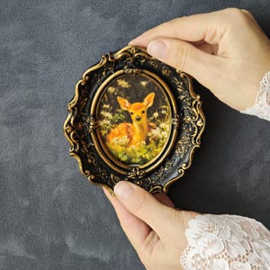 May include: An oval-shaped wall art piece featuring a small fawn resting in a field of white flowers. The artwork is encased in a decorative, ornate black frame with gold-coloured highlights along the edges and intricate floral relief details.
