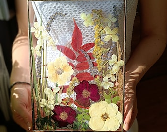 May include: A rectangular glass frame with a brass-colored metal border, filled with an arrangement of dried flowers and leaves. The botanical display includes red, yellow, and white flowers, along with green and brown foliage. The frame is held by a person.