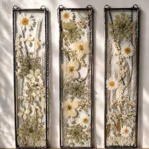 May include: Three rectangular glass panels with pressed white and yellow flowers and greenery. Each panel is framed in black metal and suspended by a chain. The floral arrangements include daisies, and other delicate blooms, creating a natural, botanical theme.