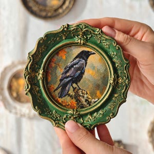 May include: A circular wall art piece featuring a detailed painting of a black crow perched on a branch against an autumn background. The artwork is encased in an ornate, scalloped green frame with gold-toned highlights along the raised edges.