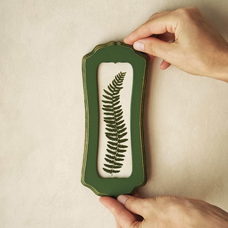 Puede incluir: Un marco de pared rectangular vertical con un acabado verde oscuro y bordes dorados. El marco presenta un borde ornamentado y festoneado, y contiene un fondo de color claro que muestra una ilustraci&oacute;n detallada de una hoja de helecho.