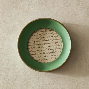 May include: A round, green picture frame with a gold-colored rim. The frame holds a piece of paper with handwritten text. The frame is set against a neutral background.