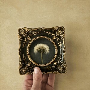 May include: A small square decorative frame with an ornate, dark brown and gold finish featuring intricate scrollwork. The frame contains a circular inset with a dark background displaying a single, preserved white dandelion seed head.