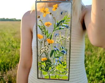 May include: A framed pressed flower art piece with yellow and blue flowers, leaves, and stems. The frame is made of metal and has a clear glass front.