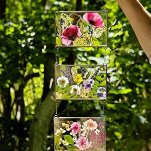May include: Three rectangular glass panels, each filled with pressed flowers, are suspended by chains. The top panel features red poppies, the middle panel has daisies and violets, and the bottom panel displays pink and white flowers. The background is a blurred green.