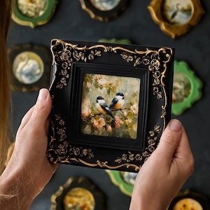 May include: A square black picture frame with ornate gold rose detailing at the corners. The frame holds a small painting of two birds perched on a branch amidst soft-toned flowers and foliage, featuring visible brushstrokes and textured paint.