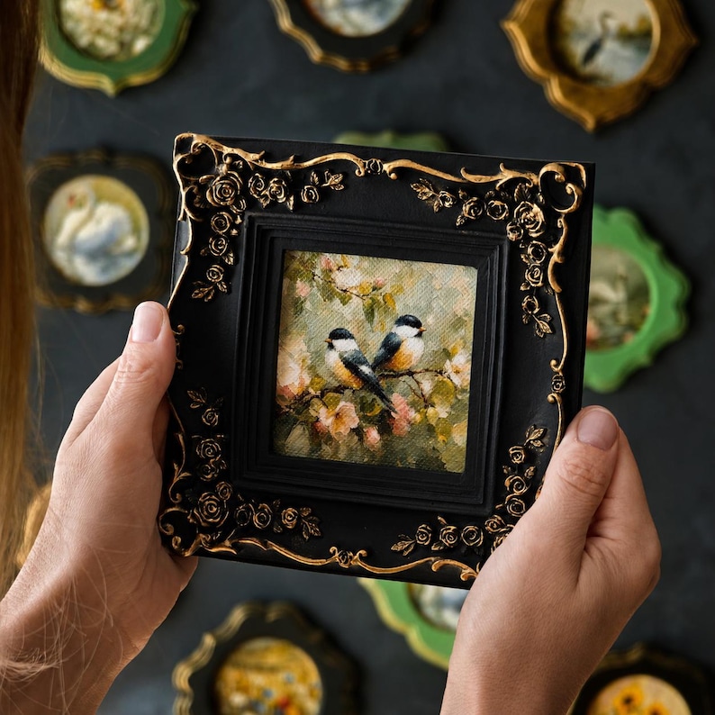 May include: A square black picture frame with ornate gold rose detailing at the corners. The frame holds a small painting of two birds perched on a branch amidst soft-toned flowers and foliage, featuring visible brushstrokes and textured paint.